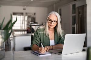 Senior stylish woman taking notes in notebook while using laptop