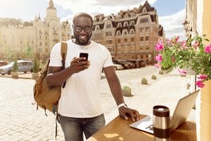 In touch. Pleasant joyful smiling man using cell phone and standing near table outside the cafe