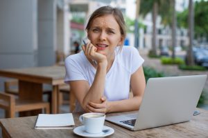 Portrait of frowning young girl sitting with laptop at cafe. Displeased young Caucasian freelancer or student wearing white T-shirt sitting at table at sidewalk cafe. Problem or difficult task concept