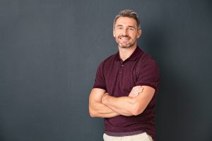 Portrait of smiling mature man standing on gray wall with folded