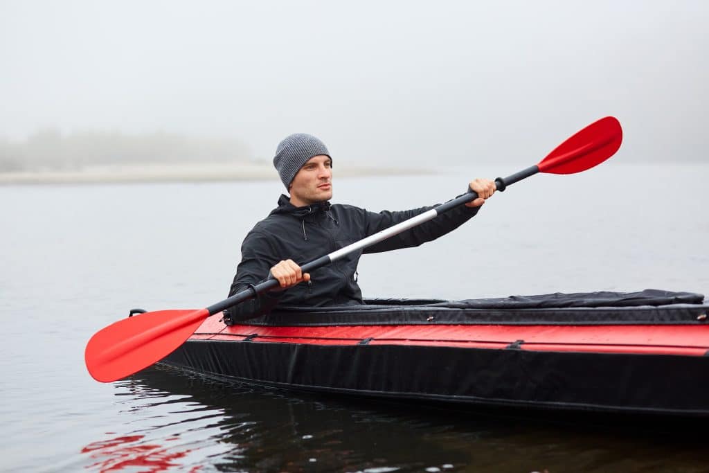 Man Paddles Red And Black Kayak With Paddle In Middle Of River O